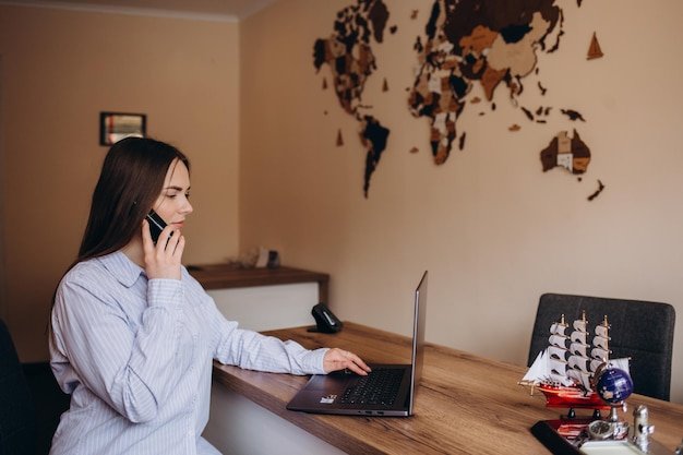 Beautiful travel agent sitting at the table in office and talking on phone