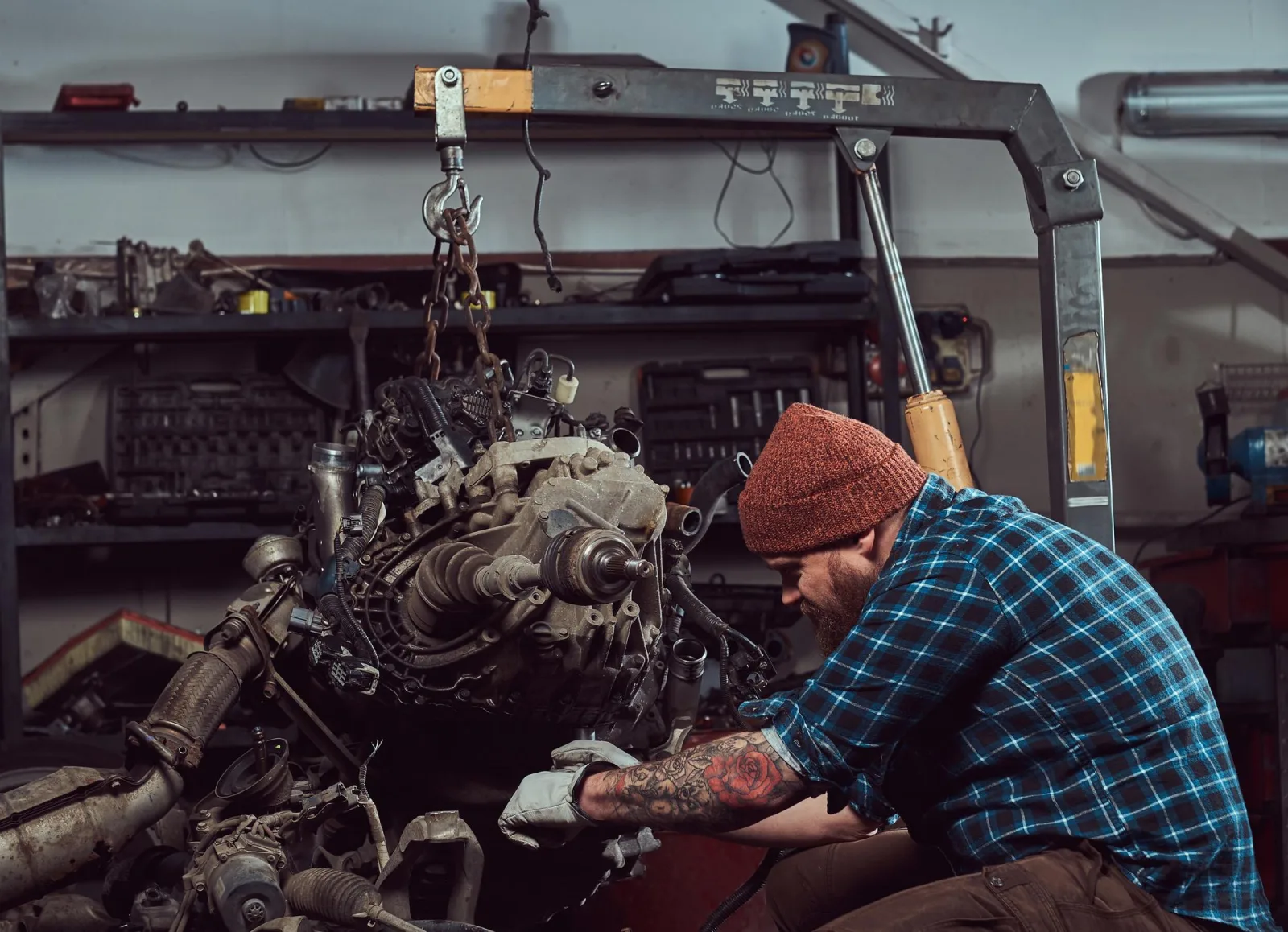 Brutal tattooed bearded mechanic specialist repairs the car engine which is raised on the hydraulic lift in the garage. Service station.