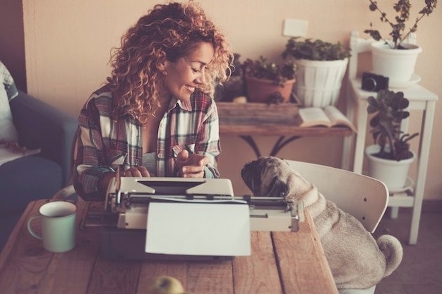 Cheerful beautiful woman with her lovely dog pug at home while write for work with old typewriter and enjoy the indoor leisure activity with cup of tea - concept of job and best friend animal