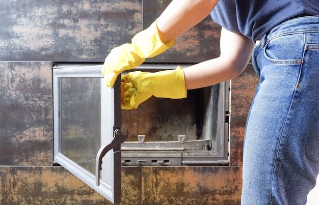 Close up of young woman in jeans yellow gloves is cleaning the fireplace Modern fireplace with glass