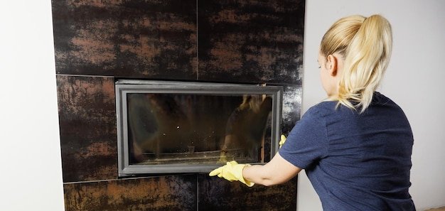 Close up of young woman in jeans yellow gloves is cleaning the fireplace Modern fireplace with glass