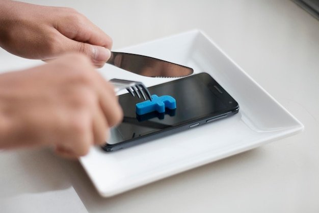 Cropped hands holding cutlery over smart phone in plate on table