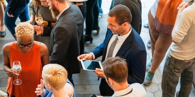 Diverse group at a social event mingling and networking People holding drinks using tablets Lively atmosphere with a mix of ethnicities and genders Diverse business people talking together