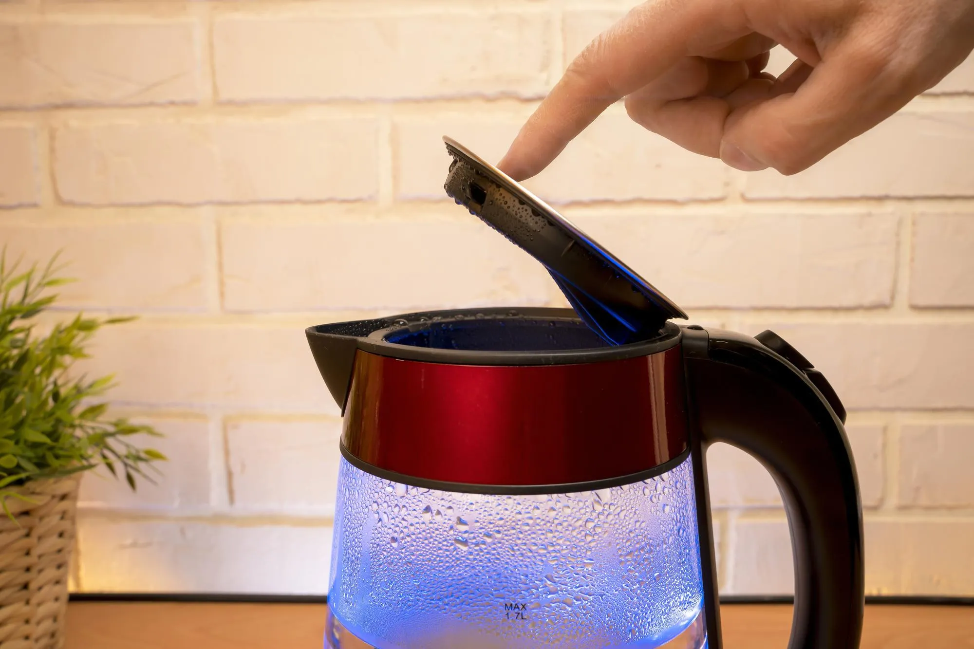 An electric kettle with boiling water and condensate on glass walls with blue neon illumination and a person 's finger closes the lid Modern Kitchen accessories