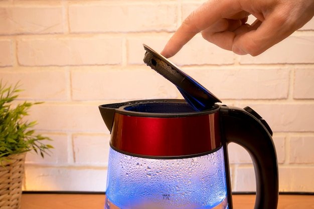 An electric kettle with boiling water and condensate on glass walls with blue neon illumination and a person 's finger closes the lid Modern Kitchen accessories