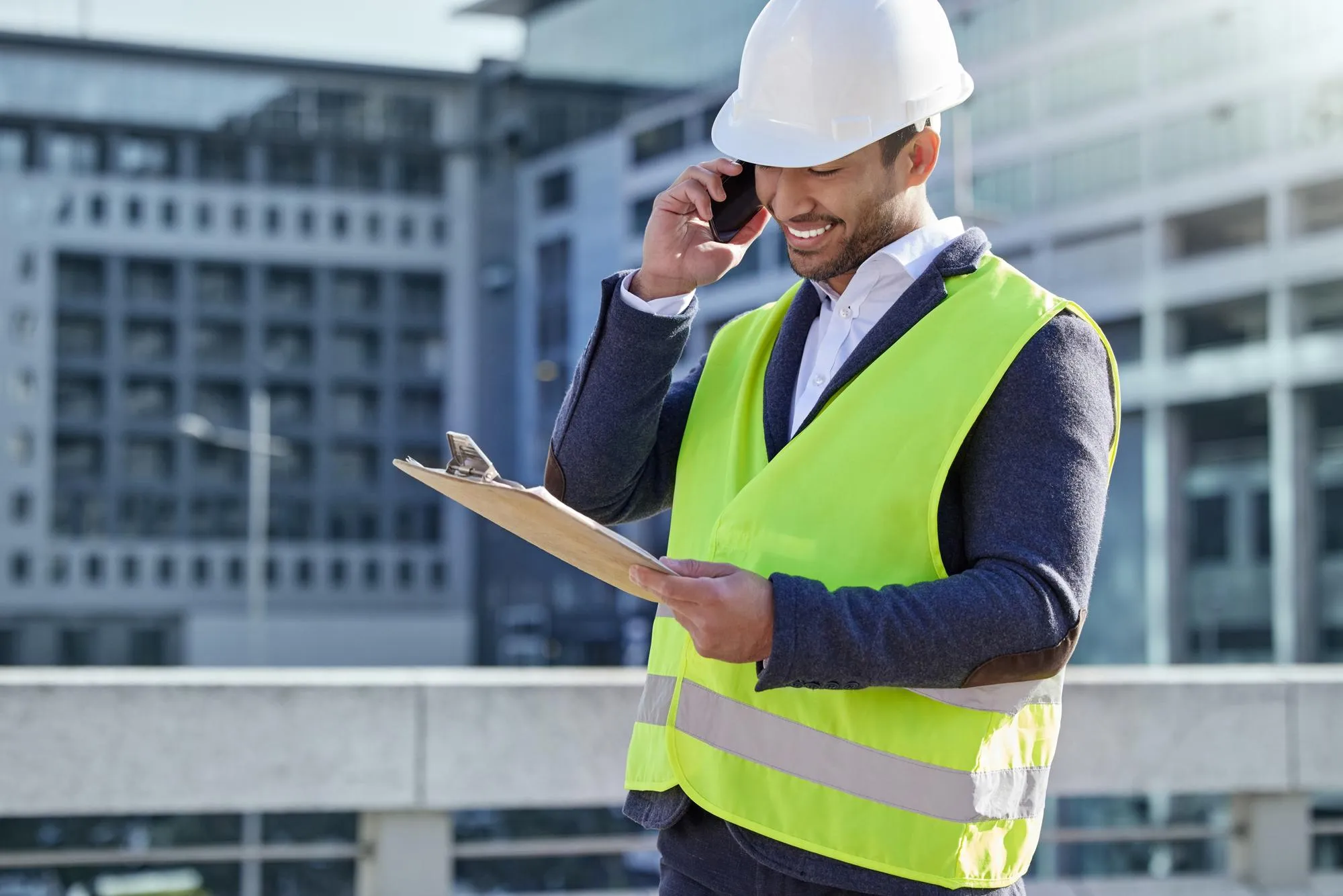 Engineer man and clipboard and call in construction site with cellphone smile and confident Male person project manager and hardhat in Abu Dhabi for building architecture and development in city