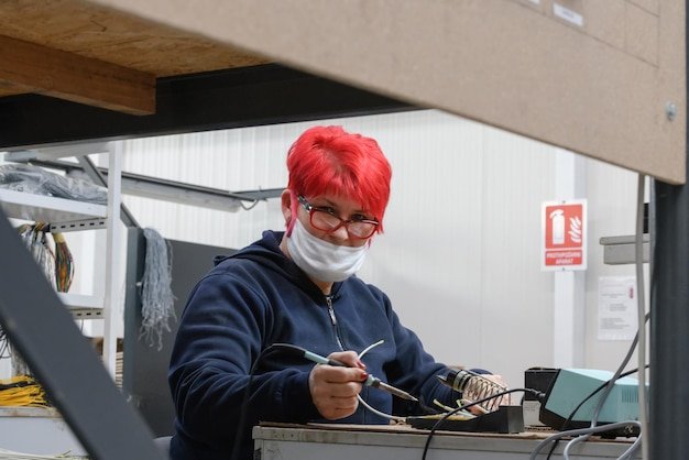 Industrial worker wearing a face mask due to a coronavirus pandemic solders cables from factory production equipment. High quality photo