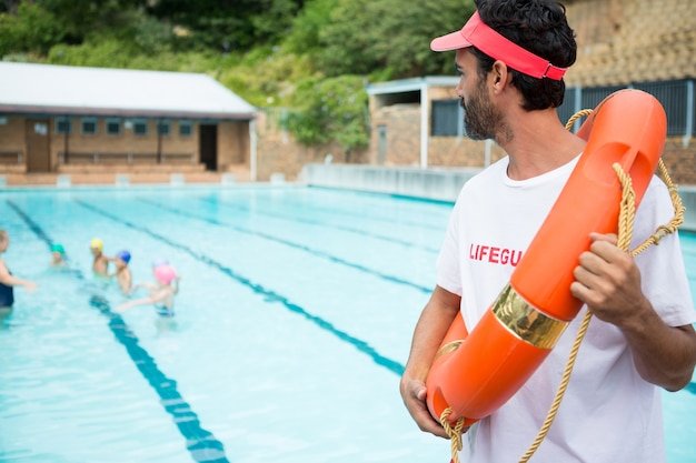Lifeguard with lifebuoy looking at students playing in the pool on a sunny day
