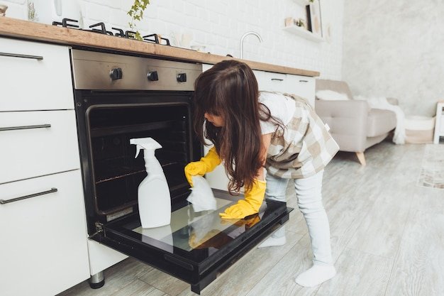 A little brunette girl wipes the oven in the kitchen at home cleaning the kitchen a child can have a stove in a white kitchen
