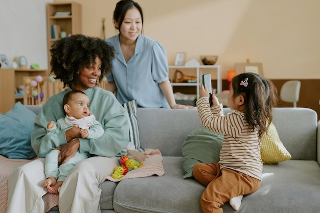 Little girl sitting on sofa and holding photo while taking photo of her mom and her african american