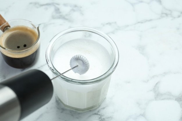 Mini mixer milk frother whipped milk in glass and coffee on white marble table closeup Space for text