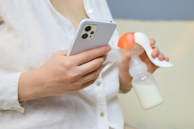 A mother holds her smartphone in one hand and a bottle of expressed milk in the other relaxed in a comfortable indoor space focusing on her device