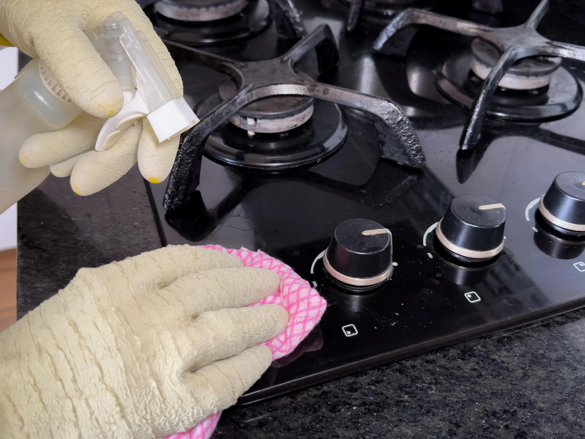 A person cleaning a modern black gas stove with a pink cloth and a spray cleaner Eliminate fat