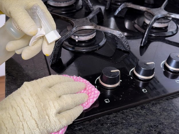 A person cleaning a modern black gas stove with a pink cloth and a spray cleaner Eliminate fat