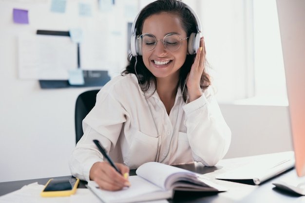 Pretty young caucasian woman in headphones dancing to music and writing lists in her planner