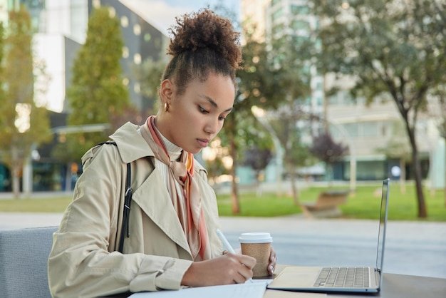 Sideways shot of serious woman dressed in coat has curly hair gathered in bun writes down notes watches webinar on portable laptop works remotely outdoors during warm sunny spring day drinks coffee