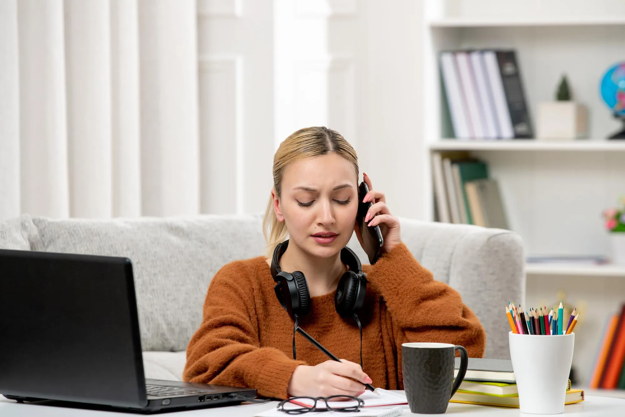 Student online cute girl in glasses and sweater studying on computer on phonecall writing notes