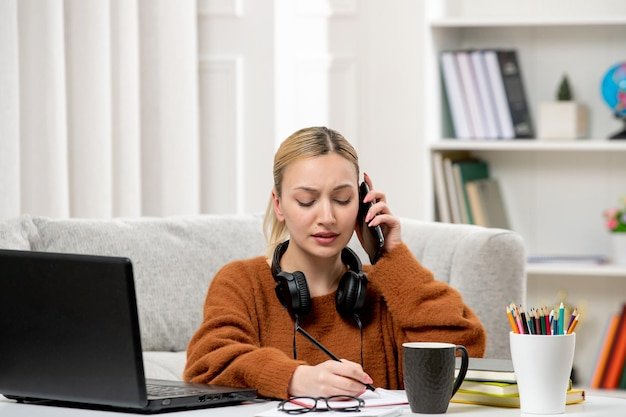Student online cute girl in glasses and sweater studying on computer on phonecall writing notes