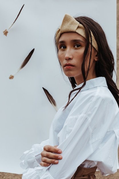 Stunning young woman with ethnic appearance posing on the white paper with feathers.