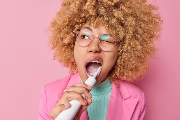 Teeth care and daily routines concept Curly haired woman brushes teeth with electric toothbrush uses mint paste cares about fresh breath and hygiene dressed formally isolated over pink wall