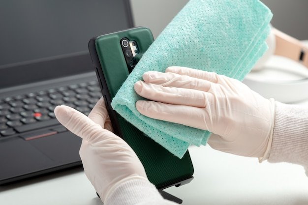Woman in gloves wipes phomelaptop with wet tissue and disinfectant during covid 19. Disinfection phomeand laptop keyboard by alcohol disinfectant by woman in mask glowes on workplace, office desk.