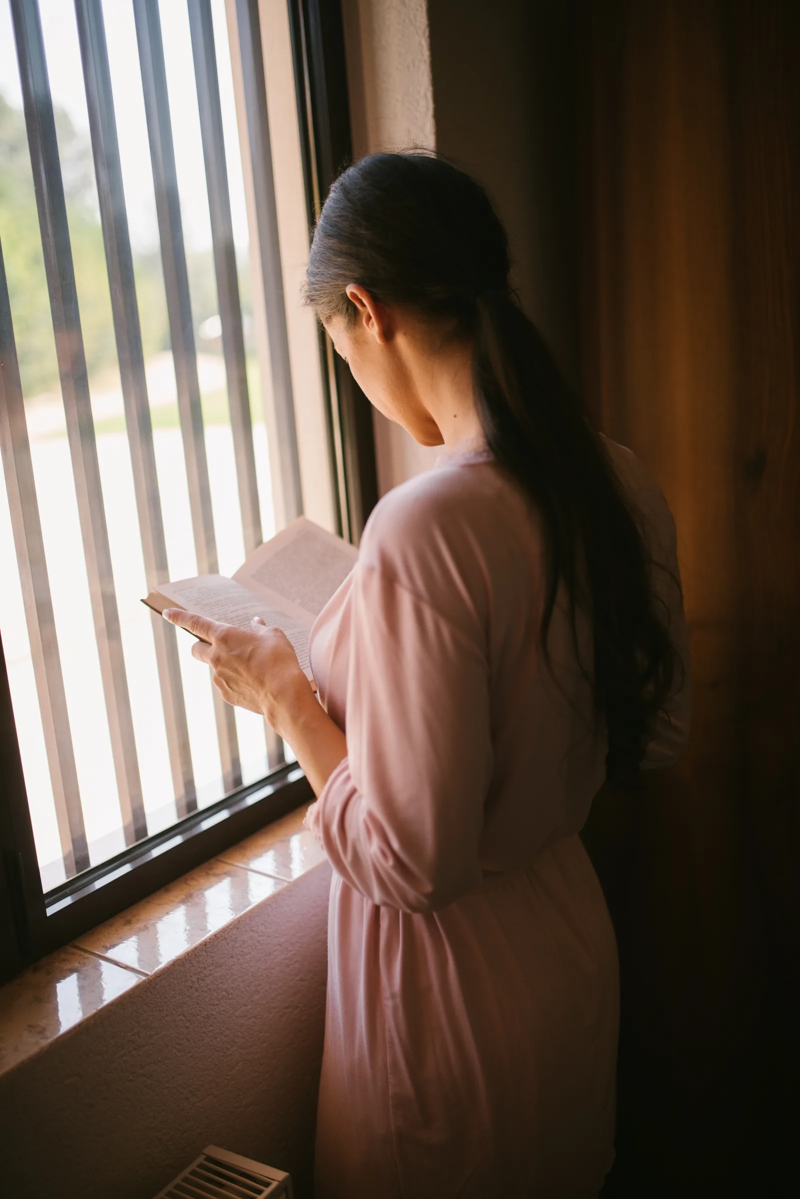 A woman stands by a window, engrossed in reading a book. Sunlight filters through the window bars, creating a warm atmosphere. She wears a soft pink robe and has long hair.