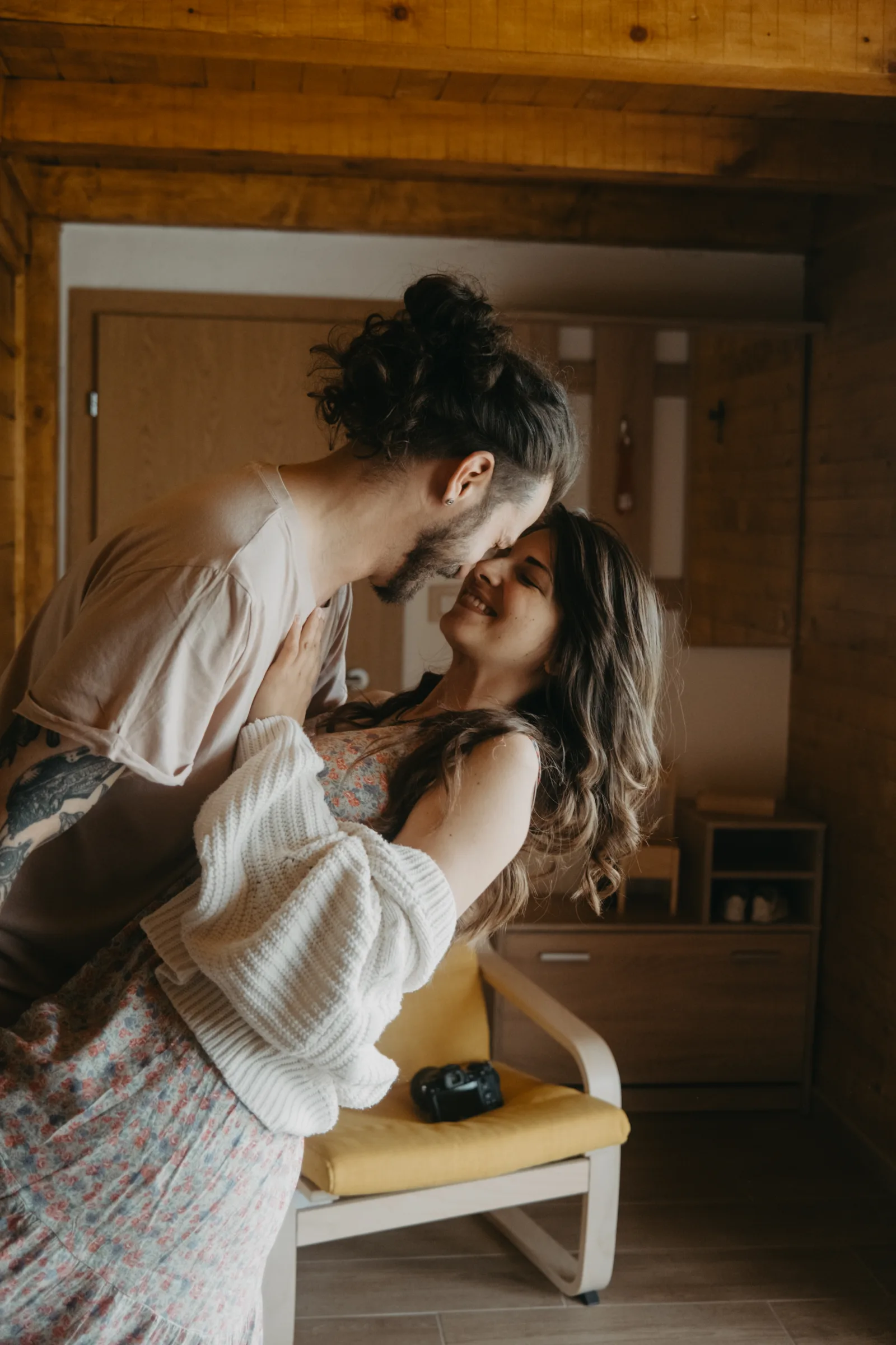 A couple smiles and holds each other close in a wooden room. They are enjoying a tender moment while standing near a yellow chair and a small table.
