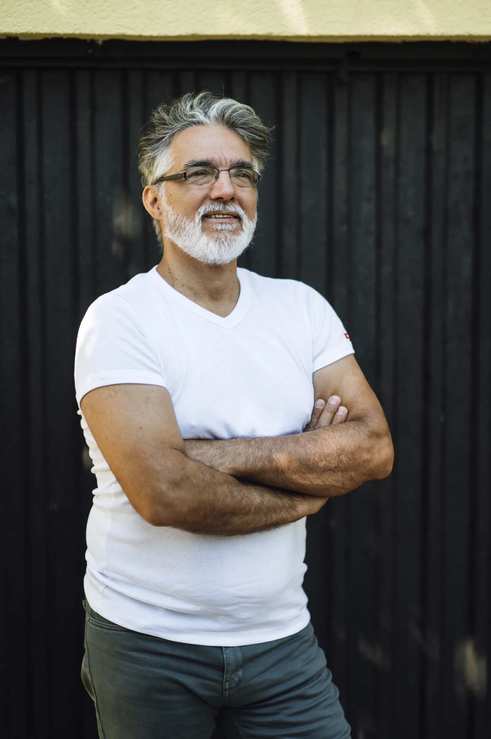 A mature man stands with arms crossed, wearing a white t-shirt and gray pants. He smiles warmly while leaning against a dark wooden wall in bright daylight.