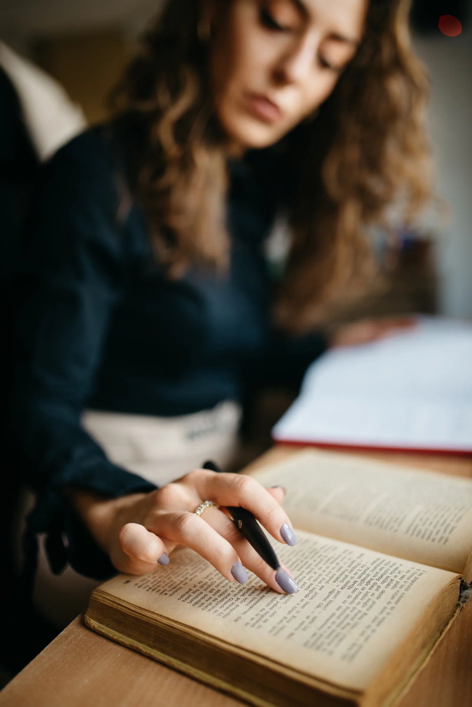 A young woman focuses on a book while using a pen to take notes at a desk in a study room. Natural light fills the space, creating a clear atmosphere for learning.