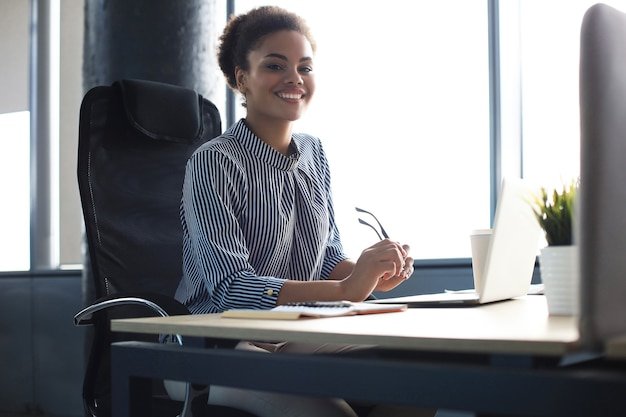 Beautiful young african american woman is sitting in the office and looking at camera.