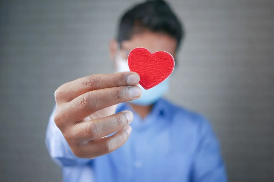 Close-up of a man with a mask holding a red heart symbol, symbolizing care and connection.