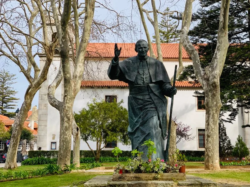 A statue of John Paul II surrounded by trees in a tranquil urban park setting.
