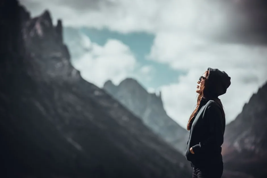 Woman in hoodie enjoying serene mountain view under cloudy sky.