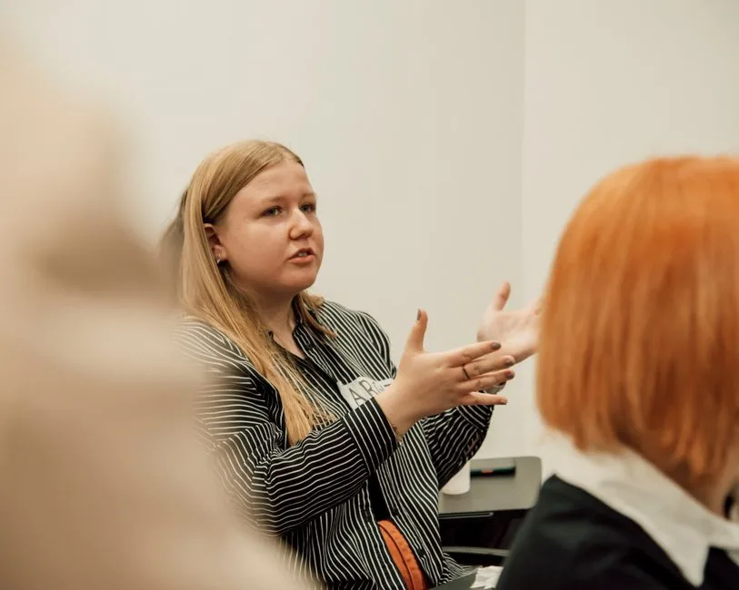 Woman with long blonde hair speaking passionately during a group meeting indoors.