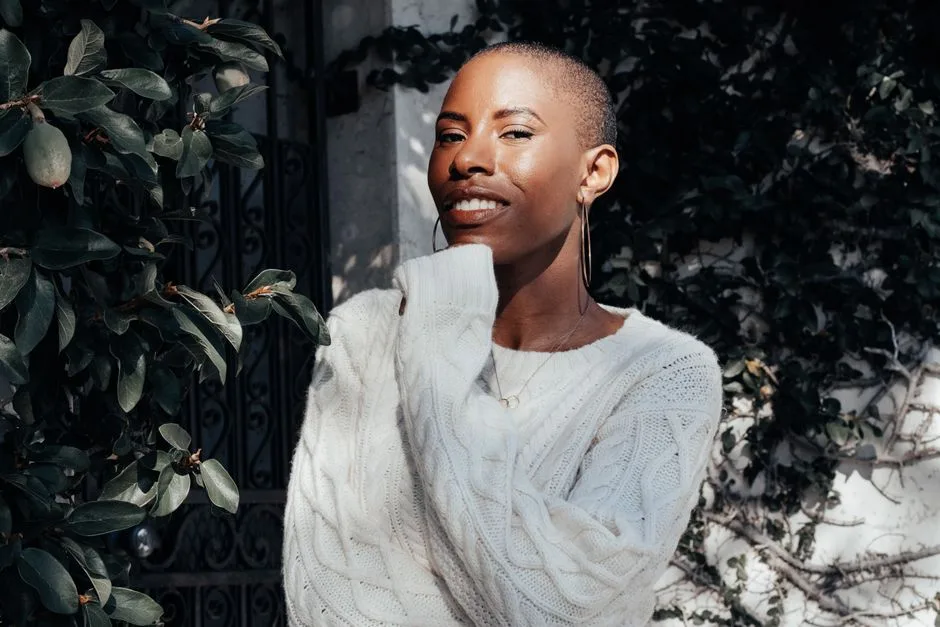 A smiling young woman in a white sweater enjoying the sunlight outdoors among green leaves.