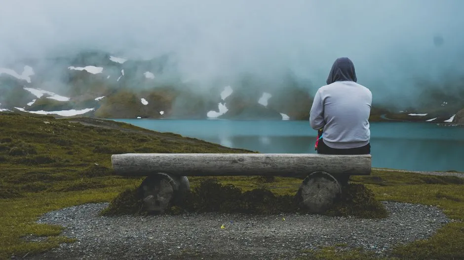 A person sits alone on a wooden bench by a misty lake, surrounded by mountains.