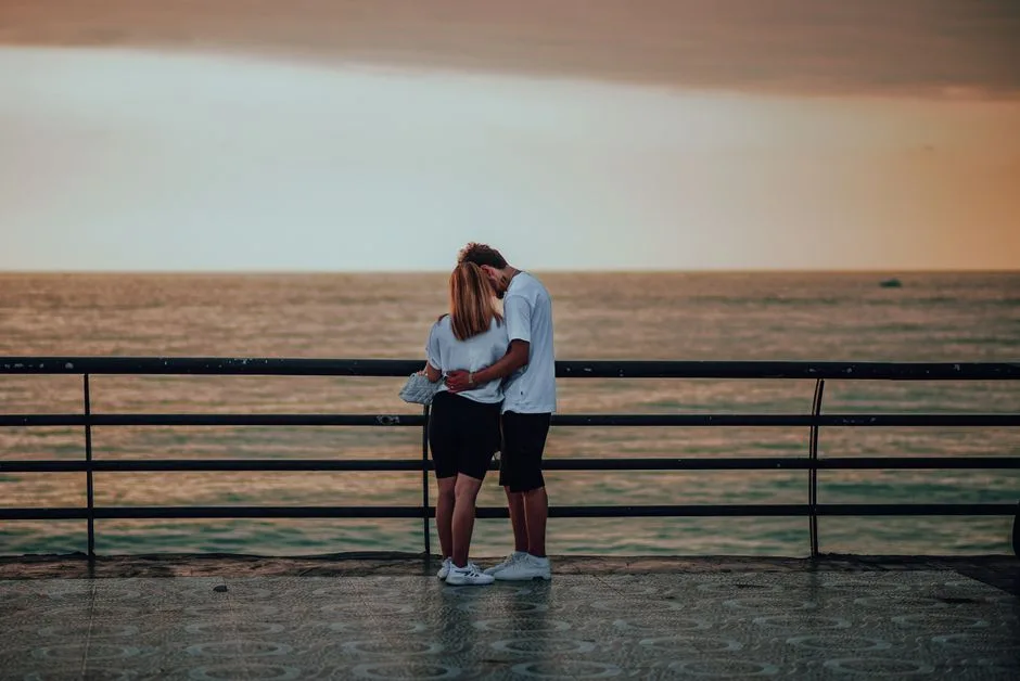 A couple embraces by the ocean at sunset, capturing a serene moment of affection.