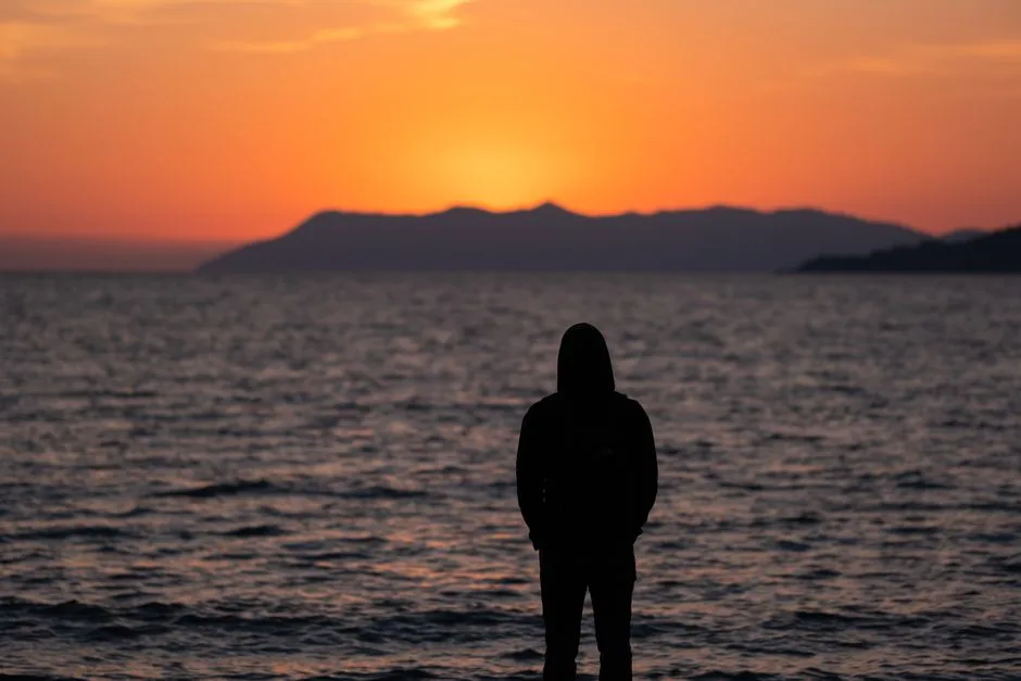 A person stands in silhouette, contemplating the sunset over the ocean horizon.