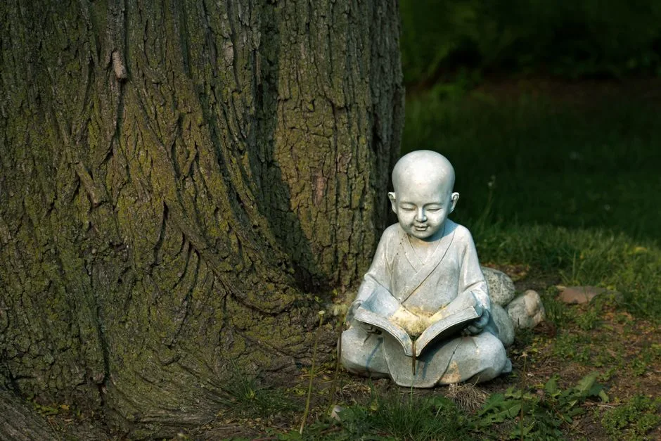 Serene Buddha statue reading by a large tree outside, symbolizing meditation and tranquility.
