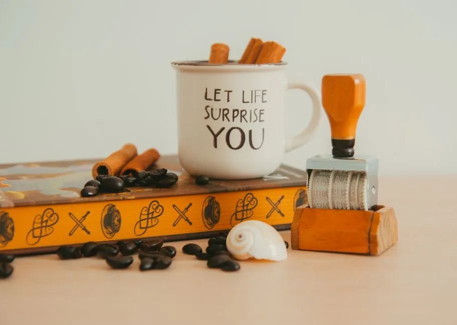 A warm scene with a mug, book, coffee beans, cinnamon sticks, date stamp, and shell.