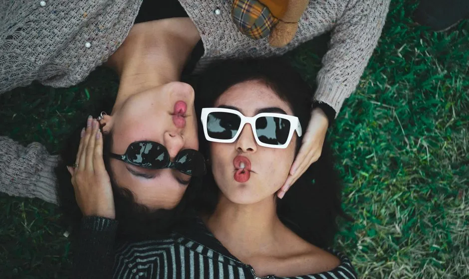 Two women lying on grass, making playful faces with sunglasses.