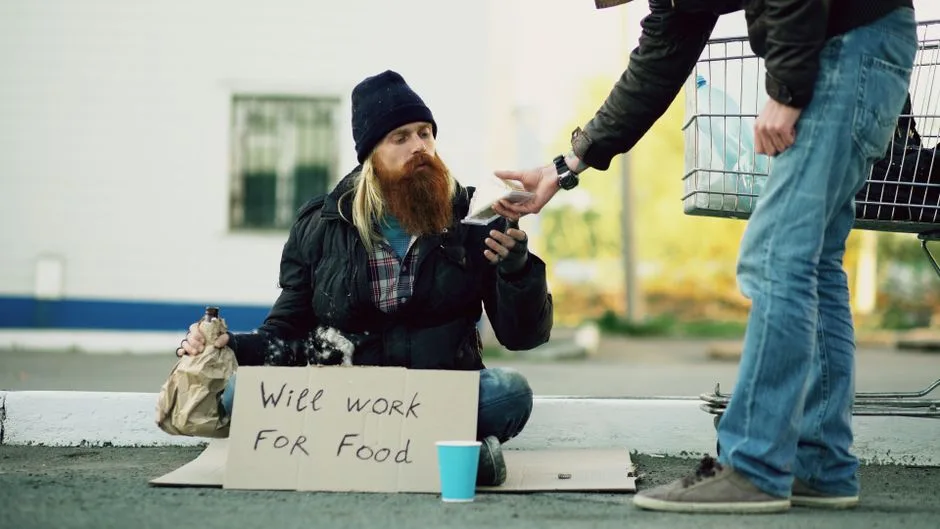A bearded homeless man receives food from a passerby outdoors.