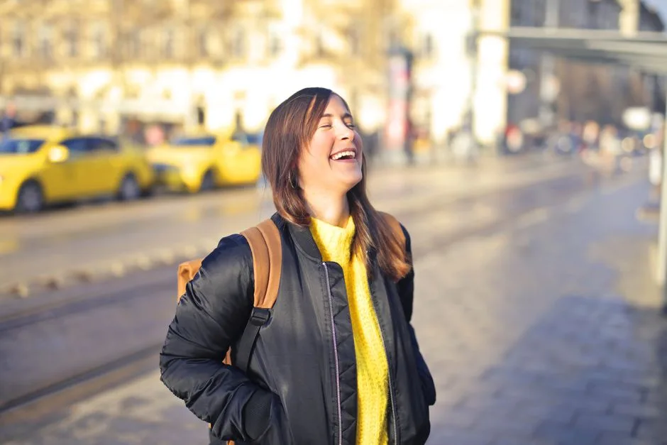 A happy woman in a black jacket laughs on a sunlit city street with yellow taxis.