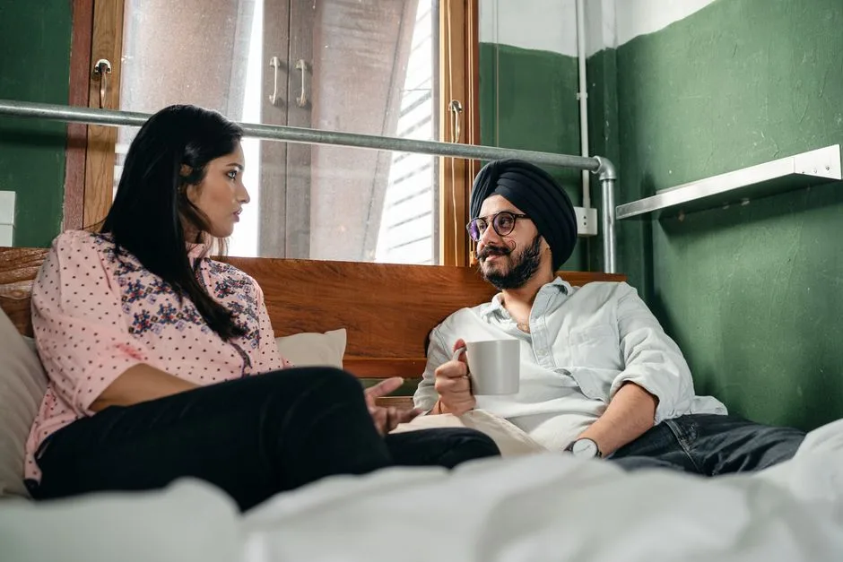 Cheerful smiling Indian male in turban and eyeglasses with cup of hot drink lying on bed with girlfriend in casual clothes and chatting