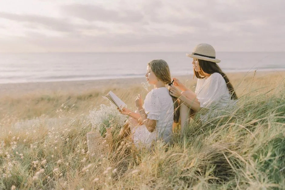 Two women enjoying a serene beach day, reading and braiding hair amidst nature.