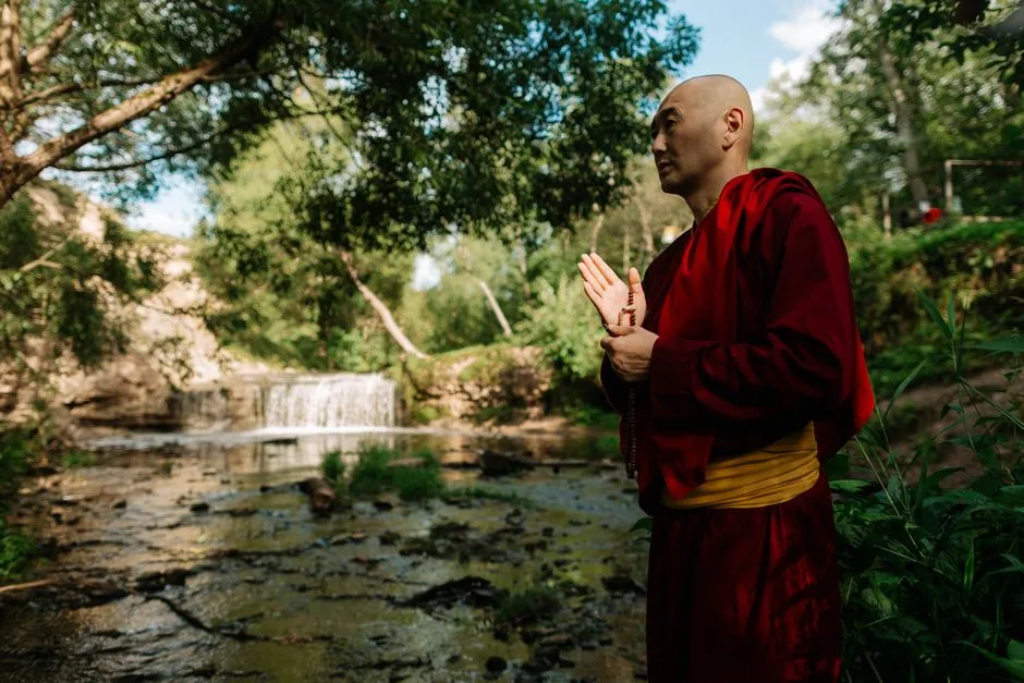 Serene scene of a Buddhist monk standing in prayer by a waterfall in nature.