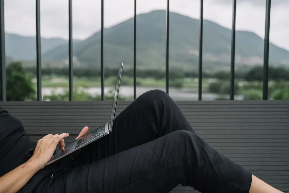 Person working remotely on a laptop, enjoying a serene mountain view from a balcony.