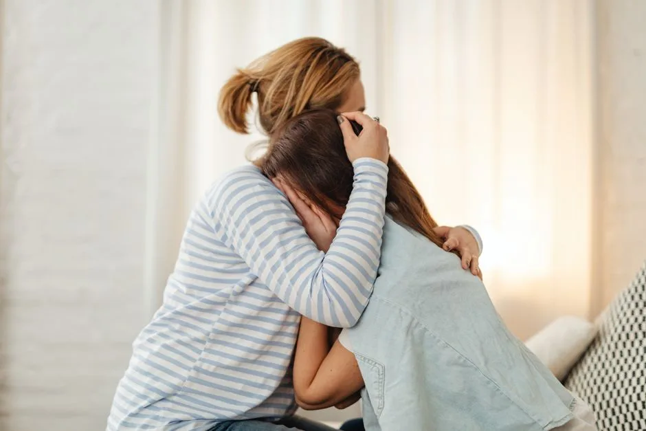 A mother lovingly comforts her teenage daughter in an emotional embrace indoors.