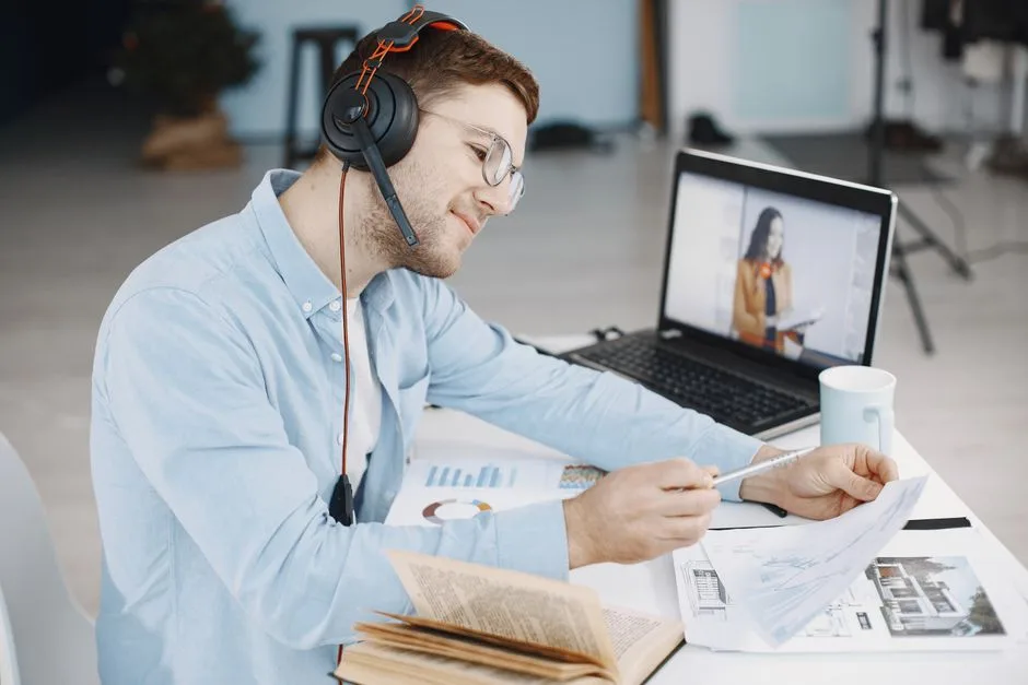 Man working remotely with headphones, reviewing documents, and video conferencing on laptop.