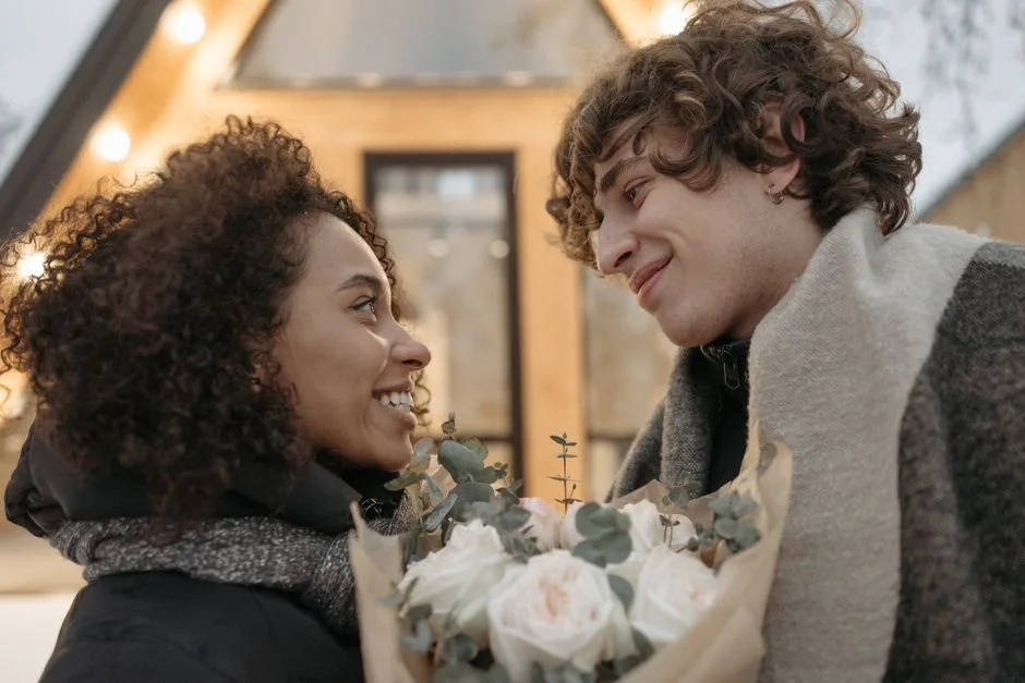 A happy couple smiling at each other with a bouquet of flowers outside a cozy house.